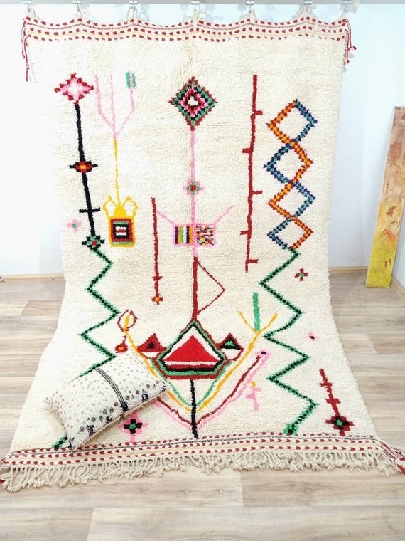 Handwoven Azilal rug with ivory wool base and colorful abstract symbols arranged in vertical lines, displayed flat to highlight texture, fringe, and traditional Moroccan craftsmanship.