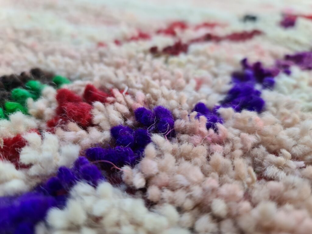 Close-up of a handwoven Moroccan rug showing thick wool pile with colorful yarn accents in purple, red, and green, highlighting texture, depth, and artisanal craftsmanship.