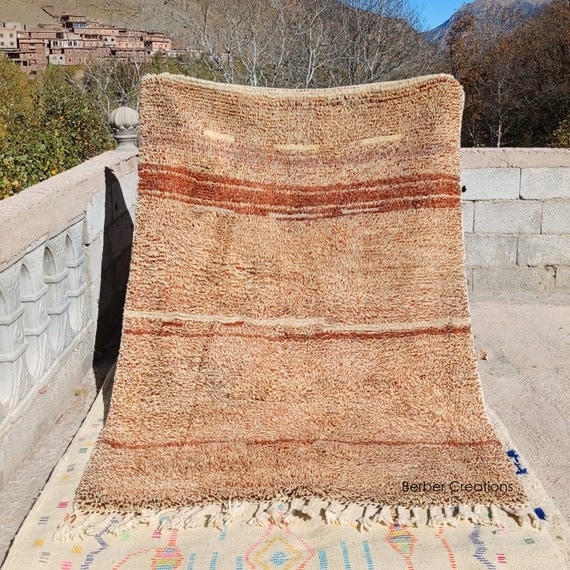Handwoven Moroccan Berber rug in warm brown and beige tones, displayed outdoors to show natural wool texture, subtle striped pattern, and traditional craftsmanship.