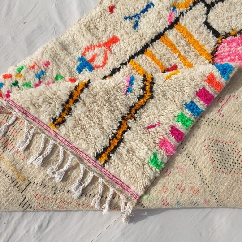 Close-up of a handwoven Moroccan Azilal rug showing thick ivory wool, colorful abstract motifs, and braided fringe, highlighting texture and traditional craftsmanship.