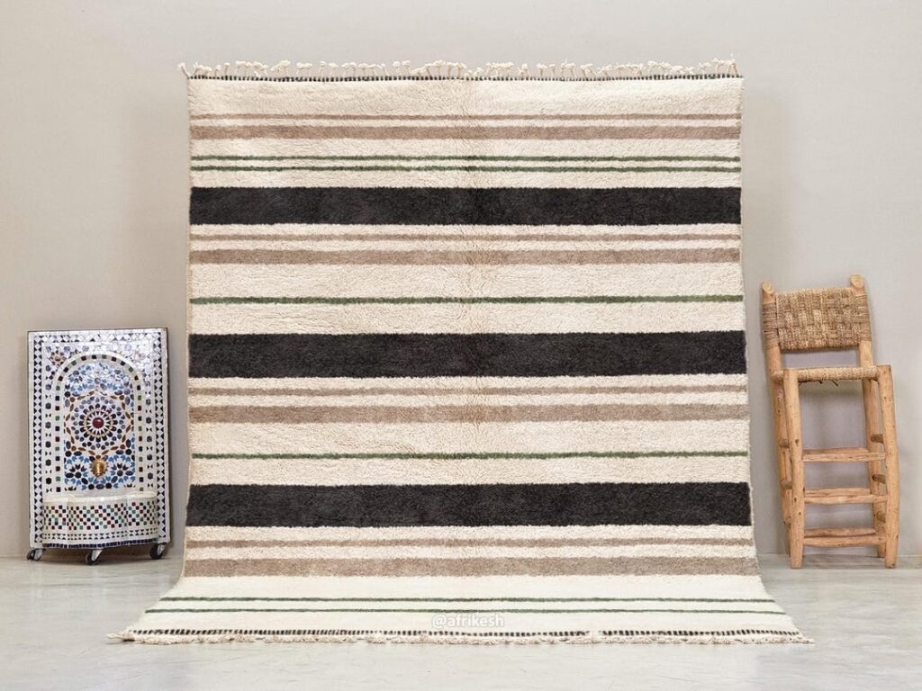 Handwoven Moroccan wool rug with horizontal stripes in ivory, beige, black, and muted green, displayed against a neutral wall.