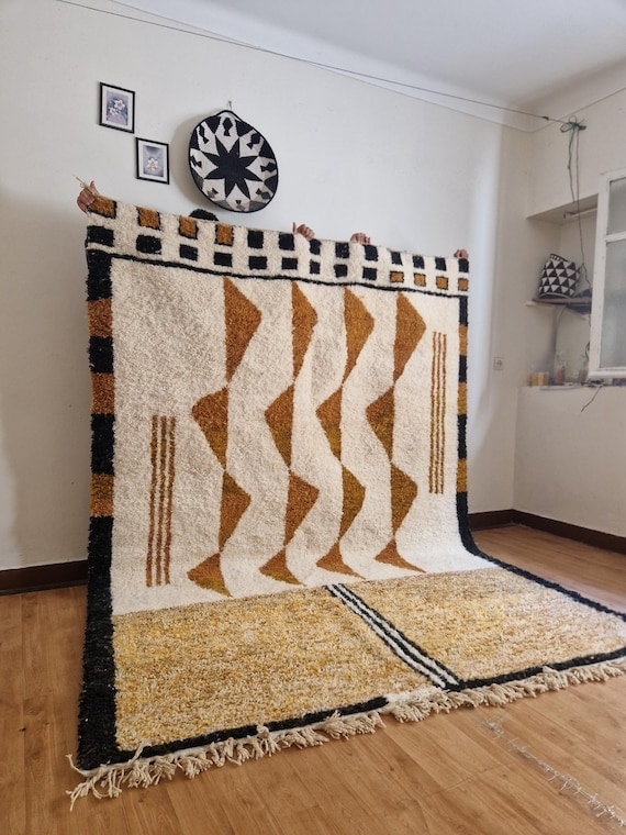 Large handwoven Beni Ourain rug featuring bold geometric motifs in ivory, black, and warm ochre tones, displayed upright in a minimalist interior.