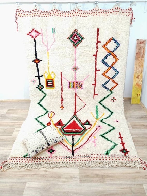 Handwoven Azilal rug with an ivory wool base and colorful abstract symbols arranged in vertical lines, displayed flat with fringe detail to showcase Moroccan craftsmanship and texture.
