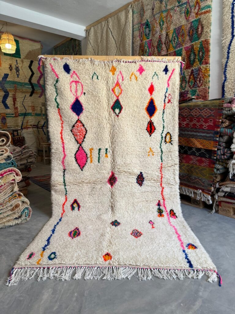 Handwoven Azilal rug with an ivory wool base and colorful abstract diamond motifs, displayed upright in a rug showroom to highlight thick pile texture, fringe, and traditional Moroccan craftsmanship.