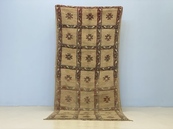 Vintage Moroccan rug with repeated geometric motifs in warm beige and brown tones, displayed flat against a neutral background to show traditional handwoven pattern and texture.