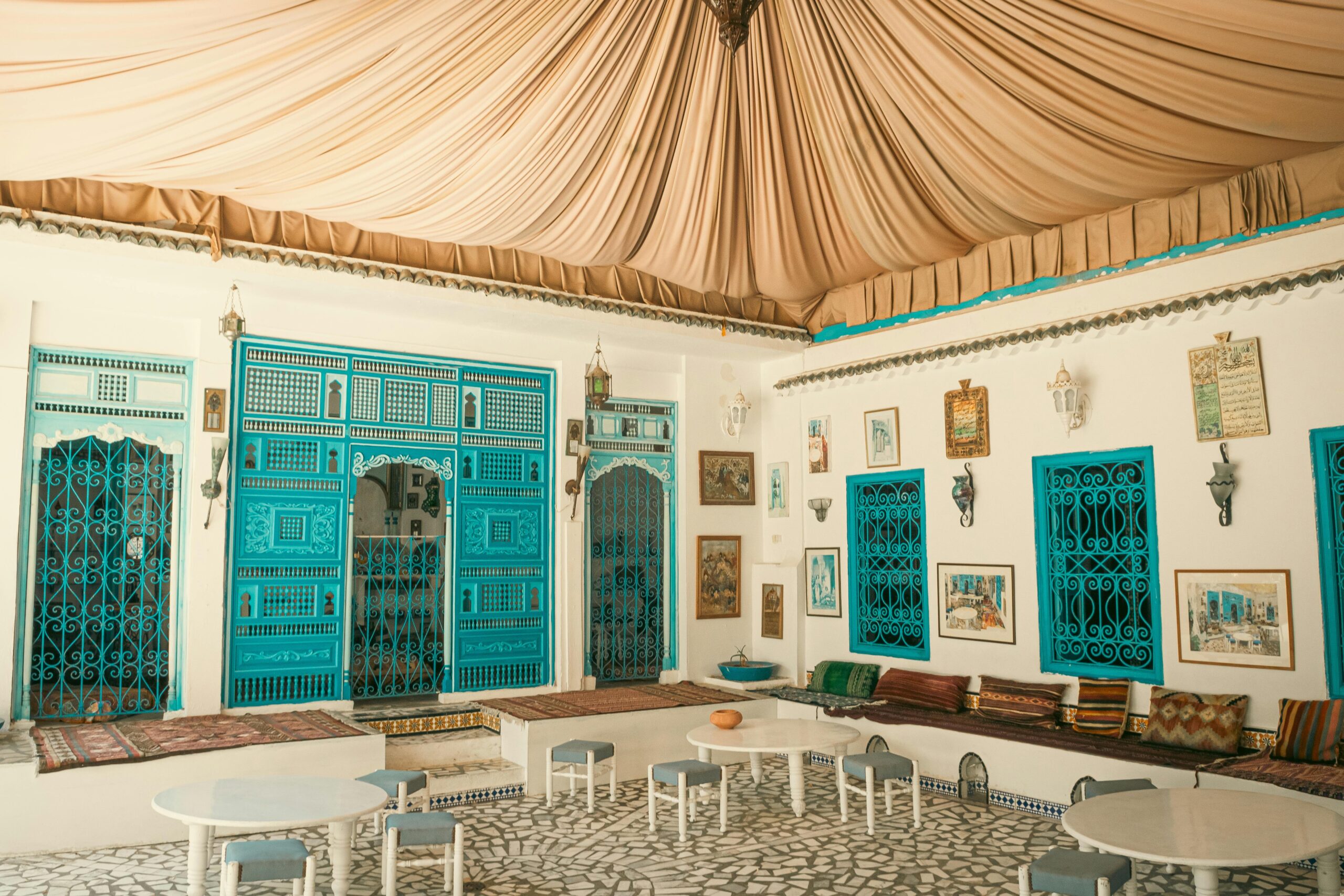 Moroccan riad living area featuring turquoise carved doors, mosaic floor tiles, low seating with colorful cushions, and draped fabric ceiling — Moroccan décor inspiration.