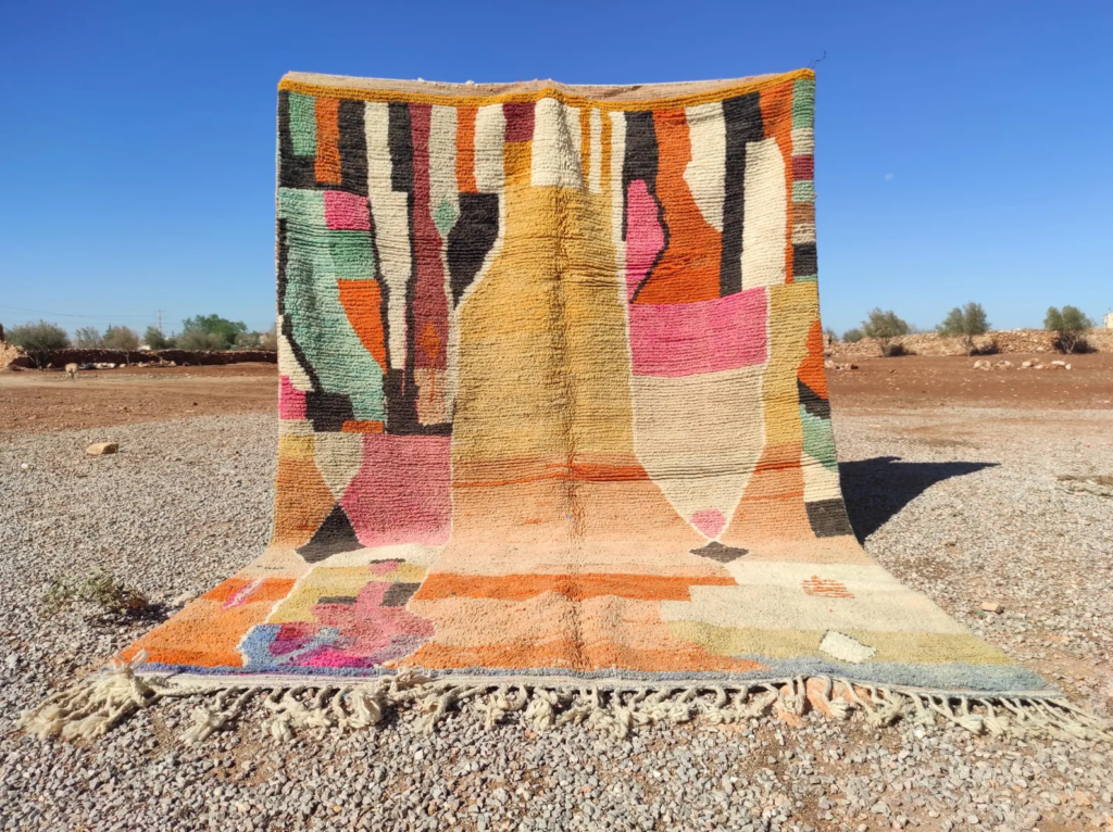 Authentic handwoven Moroccan rug displayed outdoors on desert gravel, featuring colorful geometric abstract patterns and fringe.