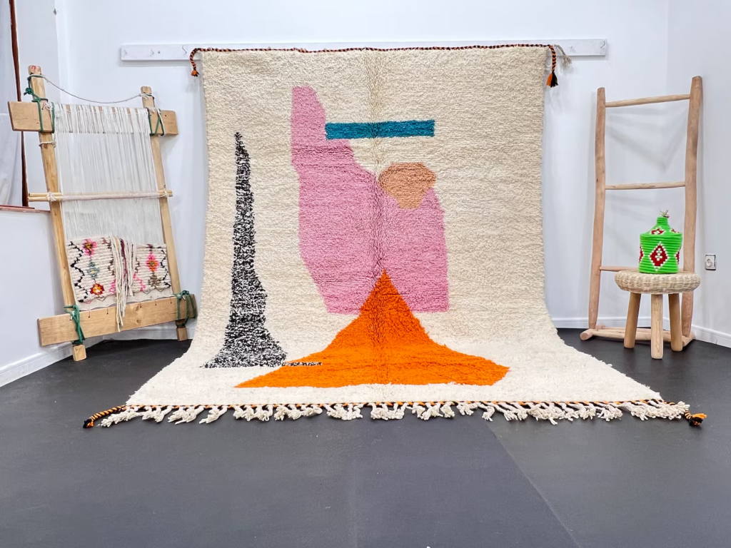 Handwoven Moroccan Berber rug with modern abstract pink, orange, black, and blue designs, displayed in a showroom beside a wooden loom and woven stool.