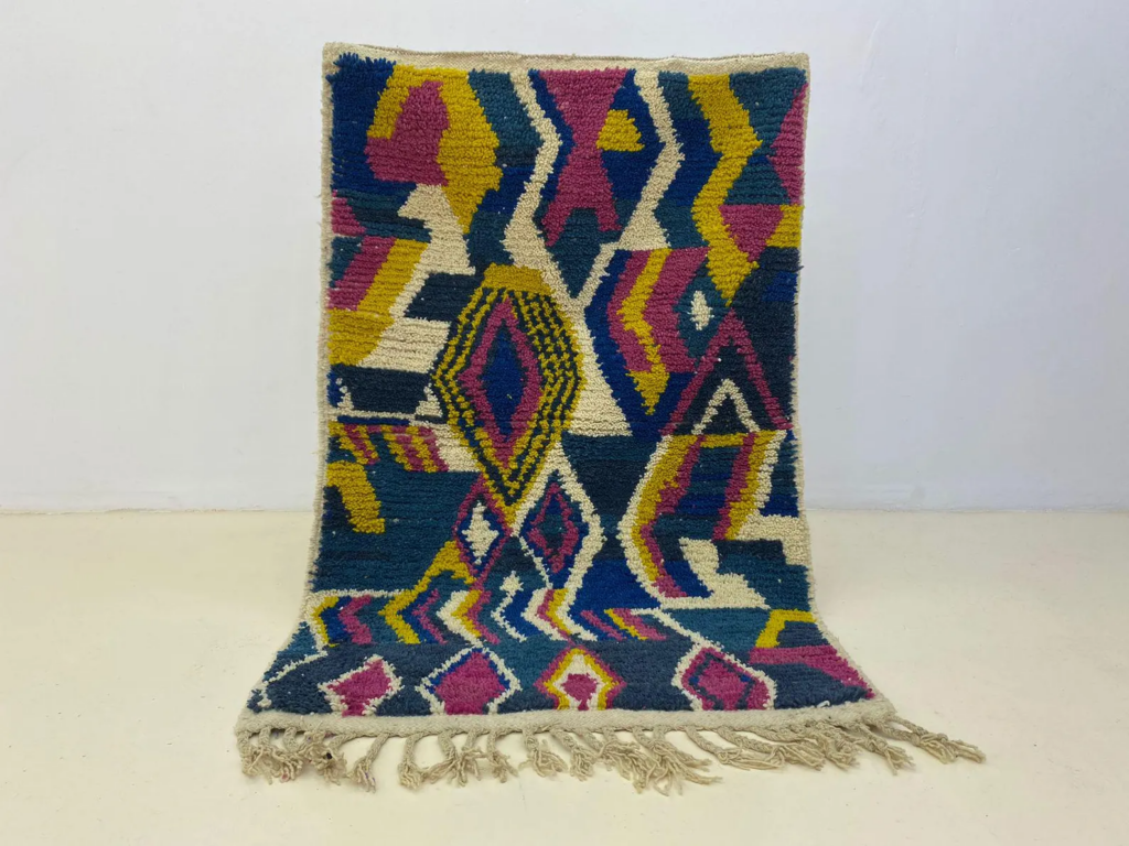 Handwoven Moroccan Azilal rug with bold geometric patterns in blue, mustard yellow, pink, and cream wool, displayed against a plain white wall.