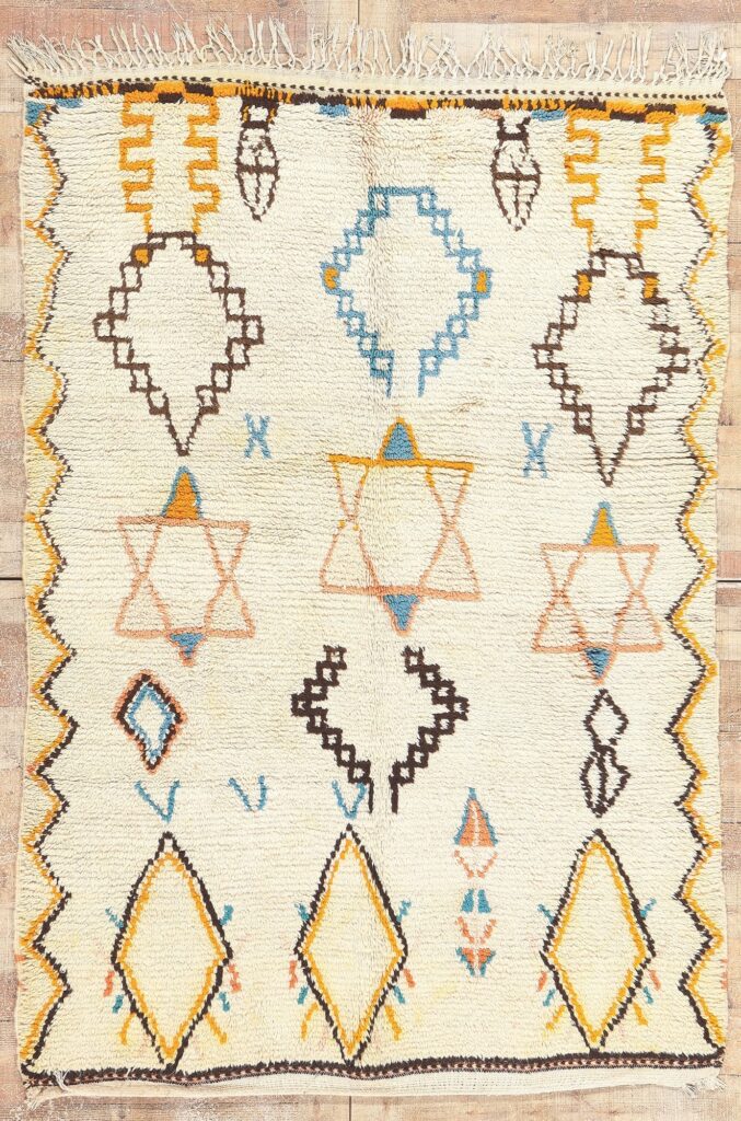 Handwoven Moroccan rug with ivory wool base and repeated geometric diamond and star motifs in soft blue, mustard, and brown tones, laid flat to highlight texture, fringe, and traditional craftsmanship.