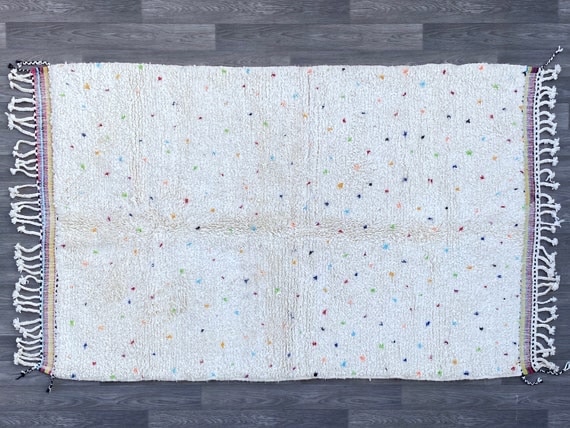 Handwoven Moroccan rug with an ivory wool base and small colorful speckled details, laid flat to show subtle texture, fringe edges, and minimalist design.